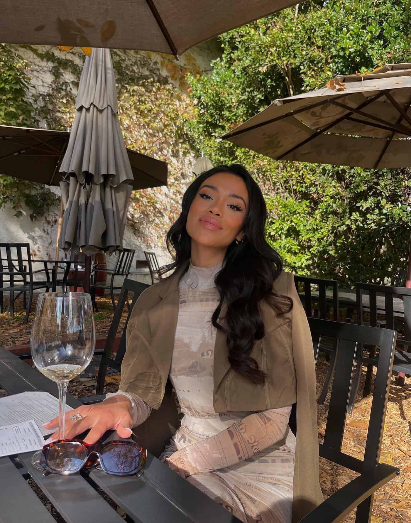woman sitting at an outdoor table drinking wine wearing a cropped jacket with a mesh long sleeve patterned dress