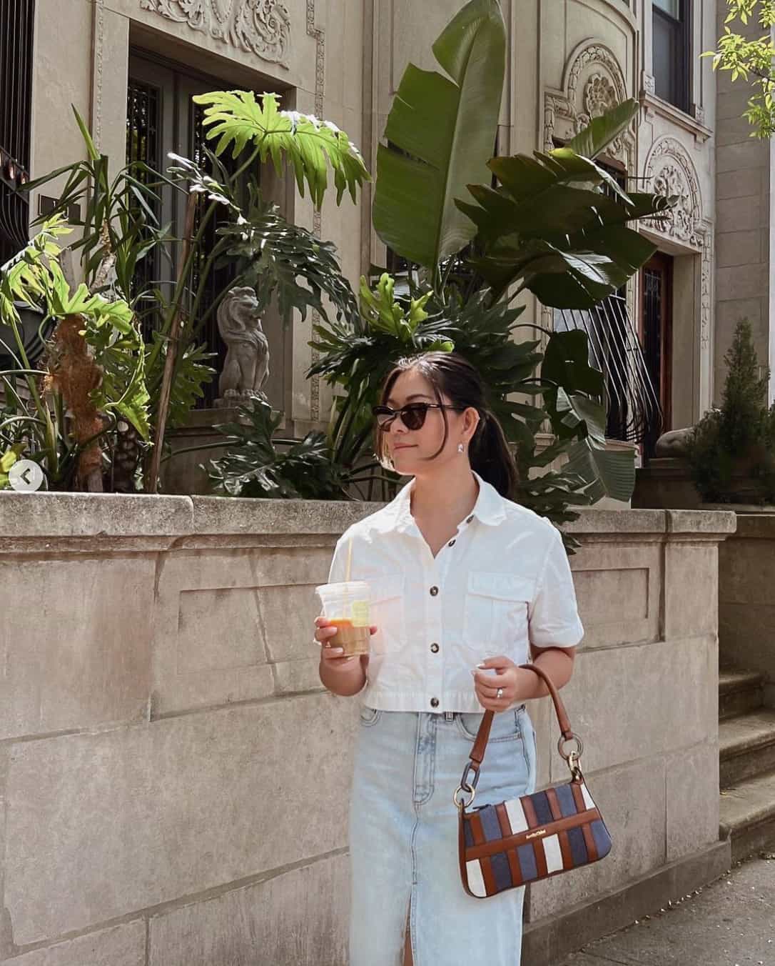 A woman wearing a white short sleeved button up with a denim midi skirt and holding a white, blue, navy, and brown leather purse from See by Chloe