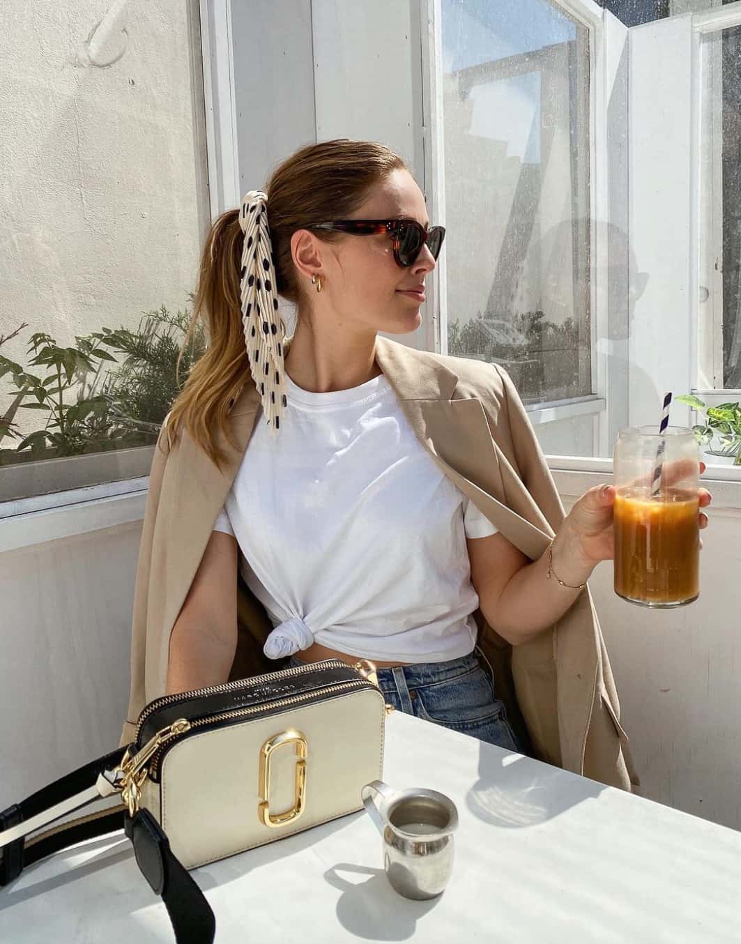 A woman wearing blue jeans, a white top, and a beige blazer with a Marc Jacobs white crossbody bag