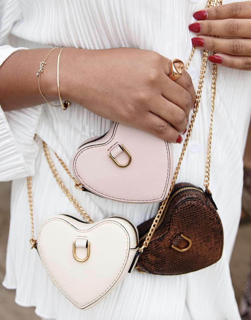 A closeup shot of a woman wearing three Fossil handbags - all shaped like hearts with gold hardware and chain straps, one white, one pastel pink, and one a textured brown leather