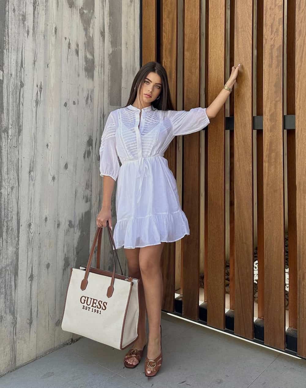 A woman holding a white canvas and brown leather Guess tote while wearing a white summery dress and brown sandals