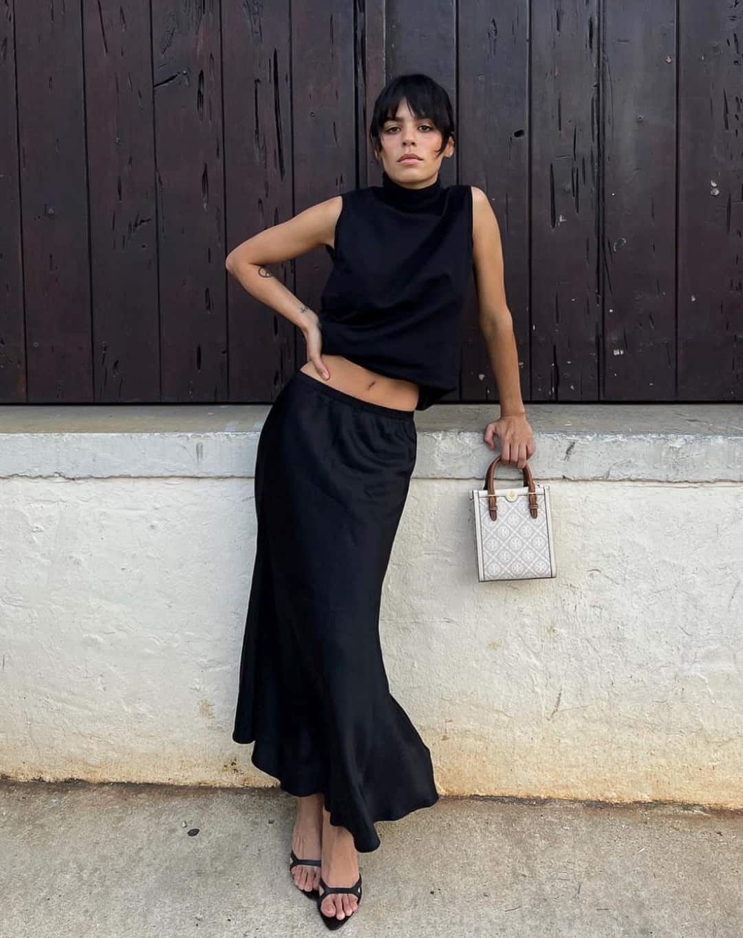 A woman wearing an all-black outfit with a midi skirt, heels, and a high-neck tank while holding a Tory Burch handbag