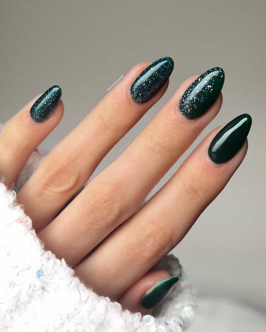 a hand with short almond nails painted a dark forest green with a glitter finish on accent nails