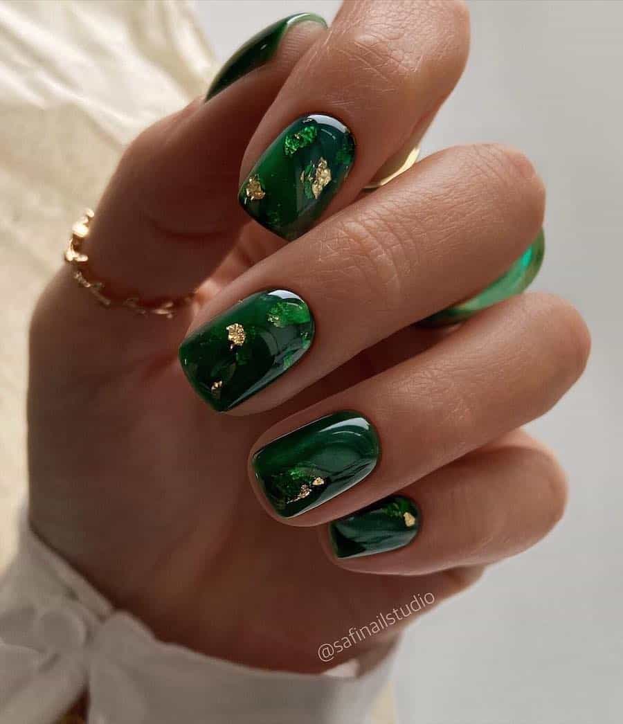 a hand with short dark green nails with metallic green and gold flake accents