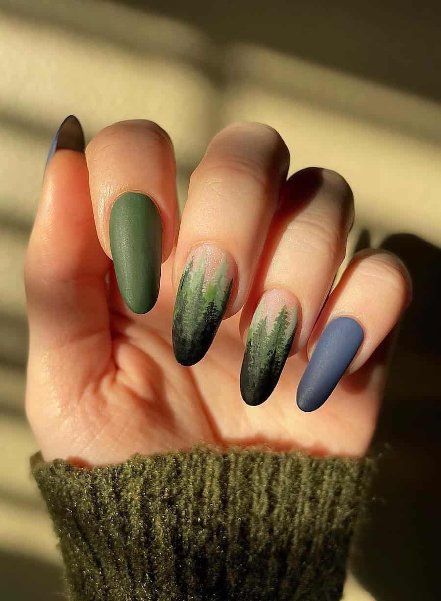 a hand with long matte almond nails painted a forest green and slate blue shade with forest nail art accent nails