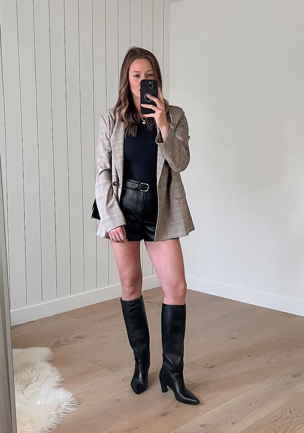 woman wearing a plaid neutral blazer over a black top with black leather shorts and black knee-high boots