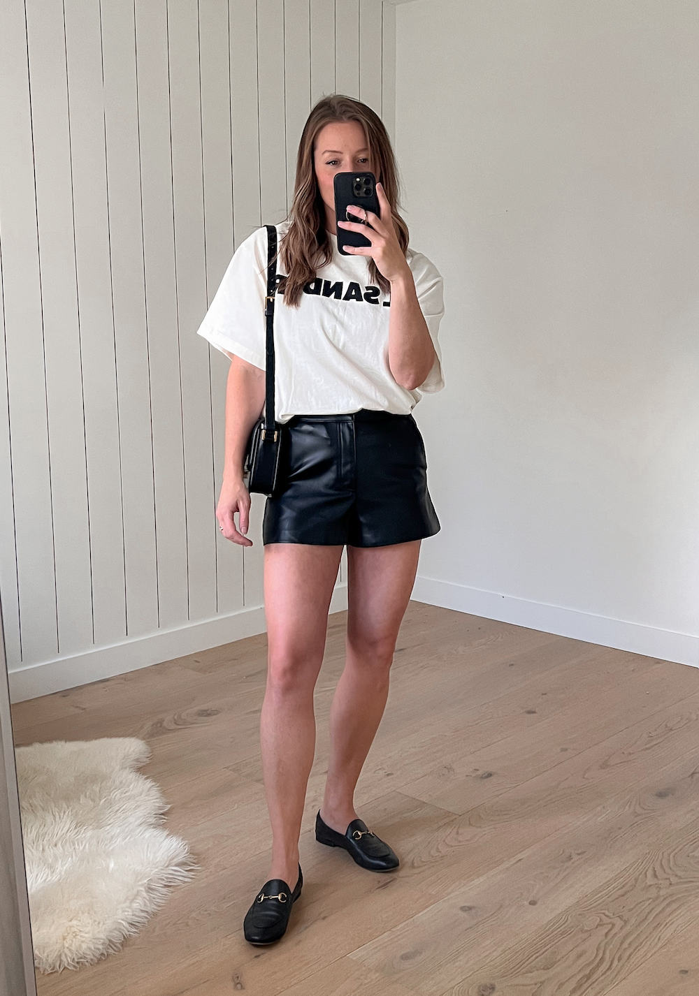 woman wearing an oversized white t-shirt with Jil Sander text and black leather shorts with black loafers