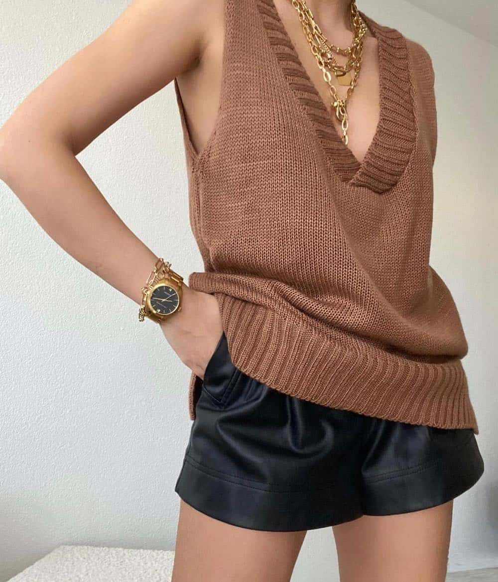 an up close shot of a woman wearing black leather shorts with a brown sweater vest, chunky gold necklaces and a gold watch and bracelets