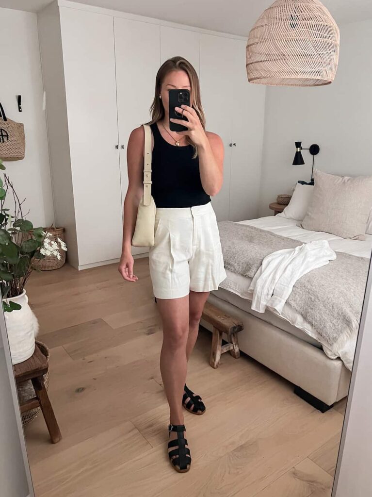 woman wearing a black tank top with ivory linen shorts, black Poppy Barley fisherman sandals, and an off-white leather shoulder bag