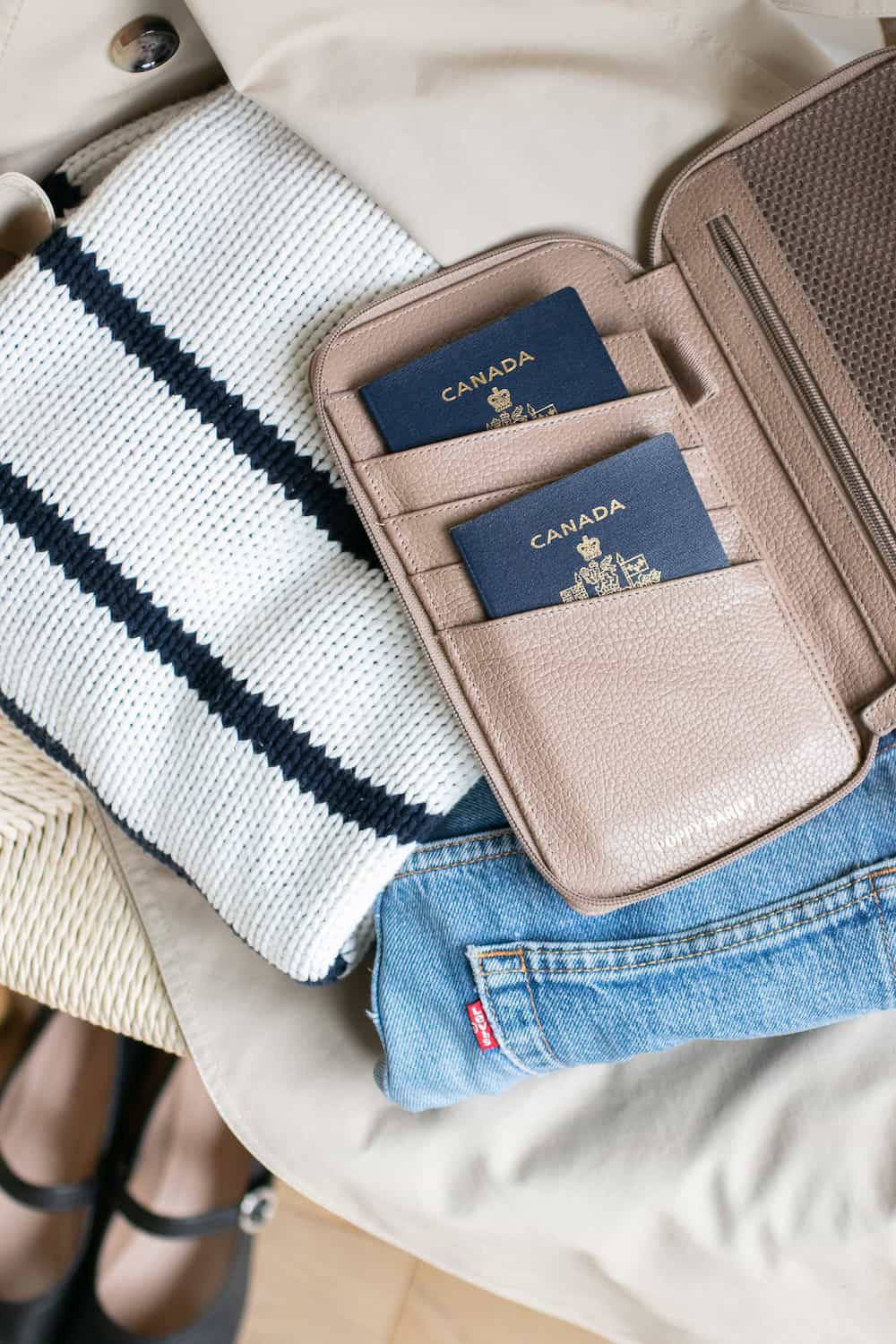 a beige leather passport holder from Poppy Barley with two Canadian passports on top of a folded striped sweater and Levis jeans