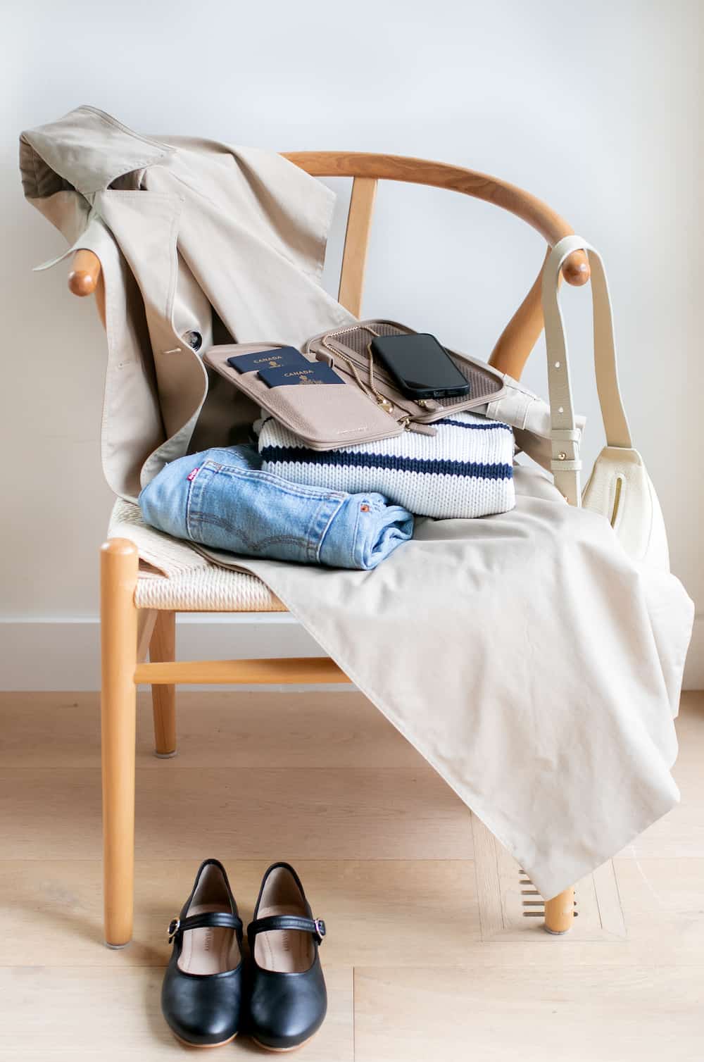 image of a wishbone chair with a trench coat slung over, a leather bag on the arm, and folded clothing with a tan passport holder on top