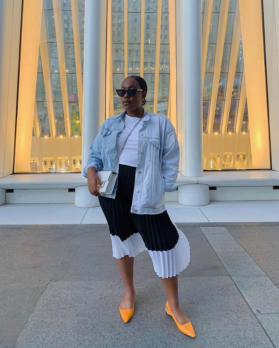 A woman wearing a black and white pleated midi skirt with a white top, a light blue denim jacket, and orange flats