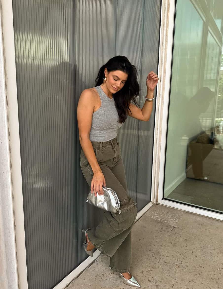 A woman wearing khaki green cargo pants, a grey tank top, silver pumps, and a silver clutch