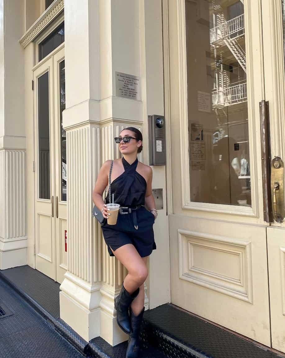 A woman wearing black shorts with a black halter top, black cowboy boots, black shoulder bags