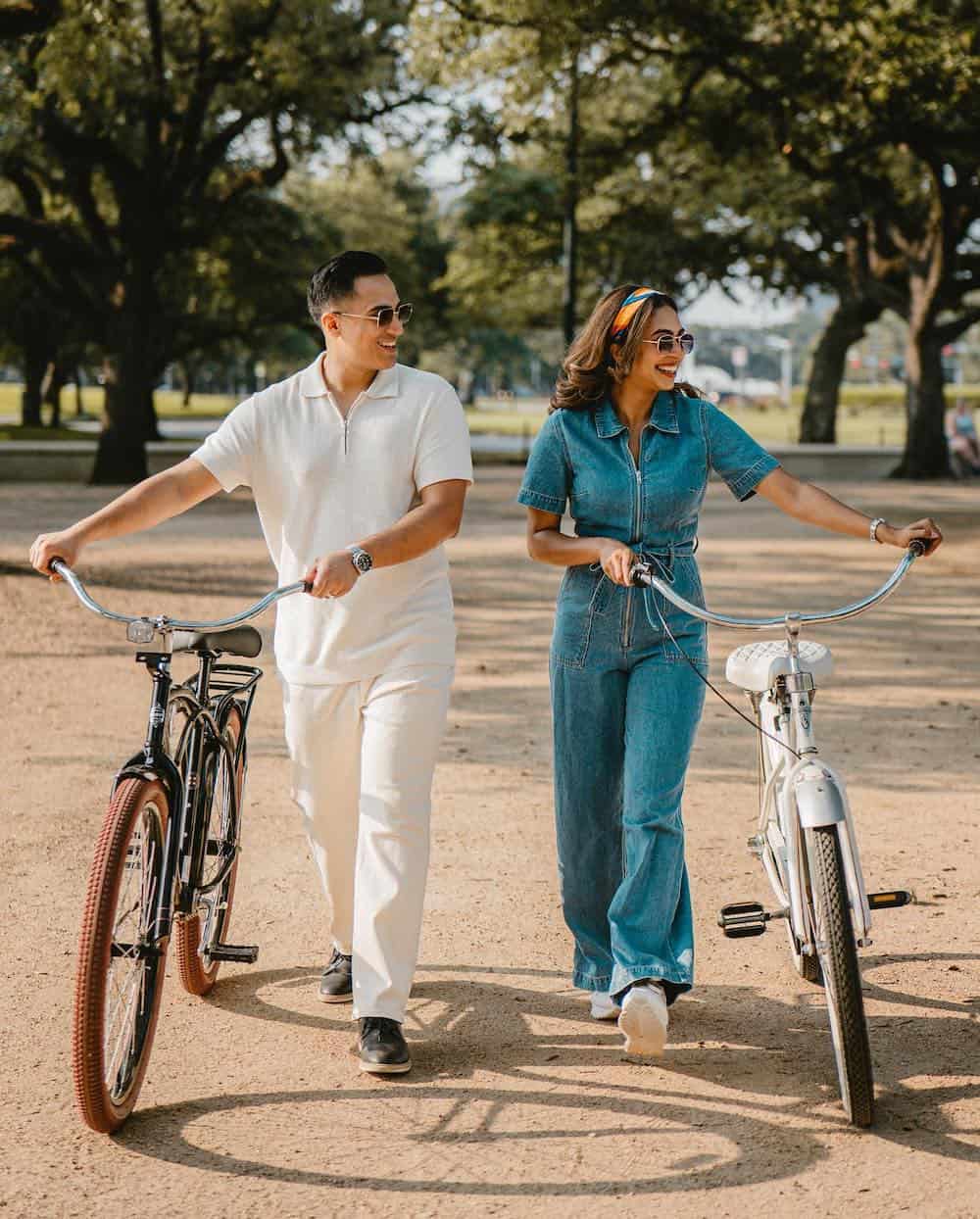 A couple walking and holding bikes side by side, the woman wearing a blue denim jumpsuit and sneakers while the man wears white pants and a white collared tee with black sneakers