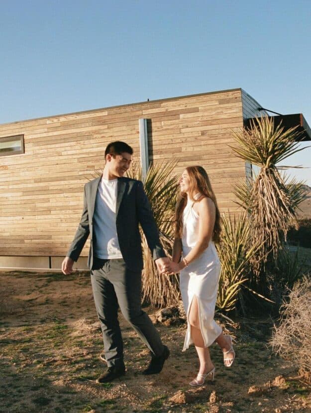 A couple's engagement photoshoot and the woman is wearing a slitted white halter top dress with white strappy heels, and the man is wearing matching black pants and blazer with a white tee
