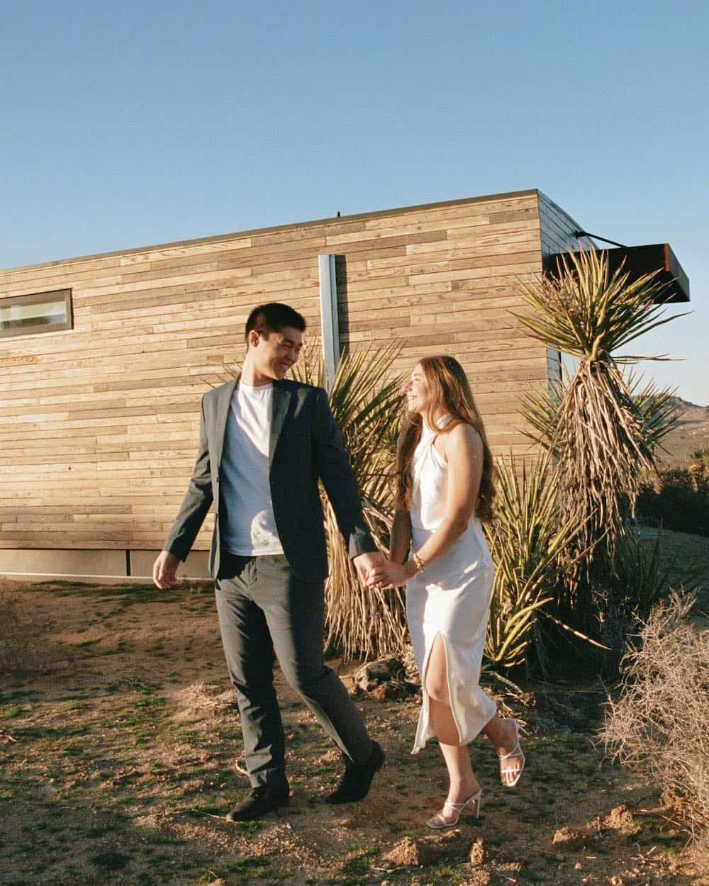 A couple's engagement photoshoot and the woman is wearing a slitted white halter top dress with white strappy heels, and the man is wearing matching black pants and blazer with a white tee