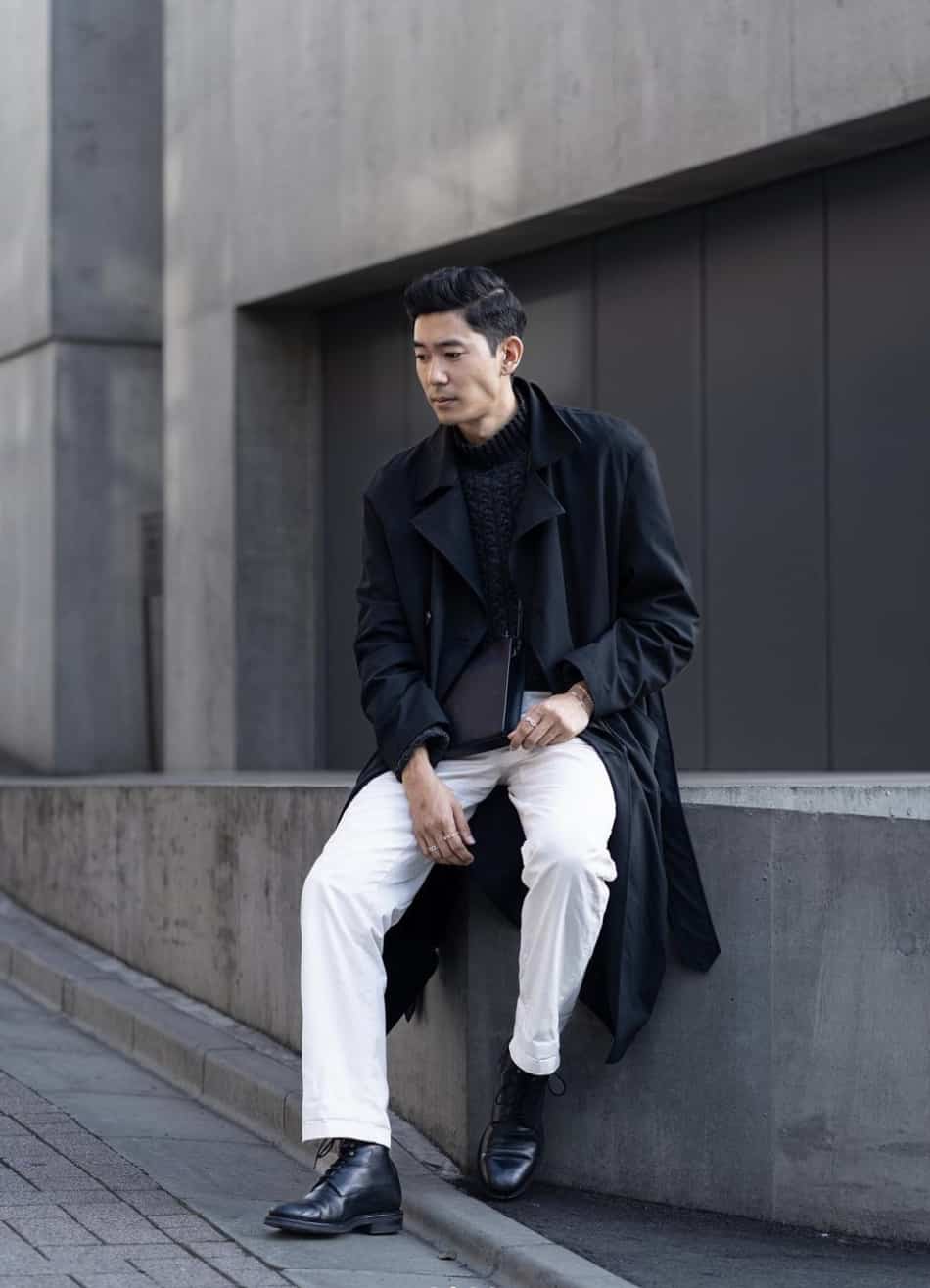 a man wearing white pants with a black mock neck sweater, a black trench coat, and black dress shoes