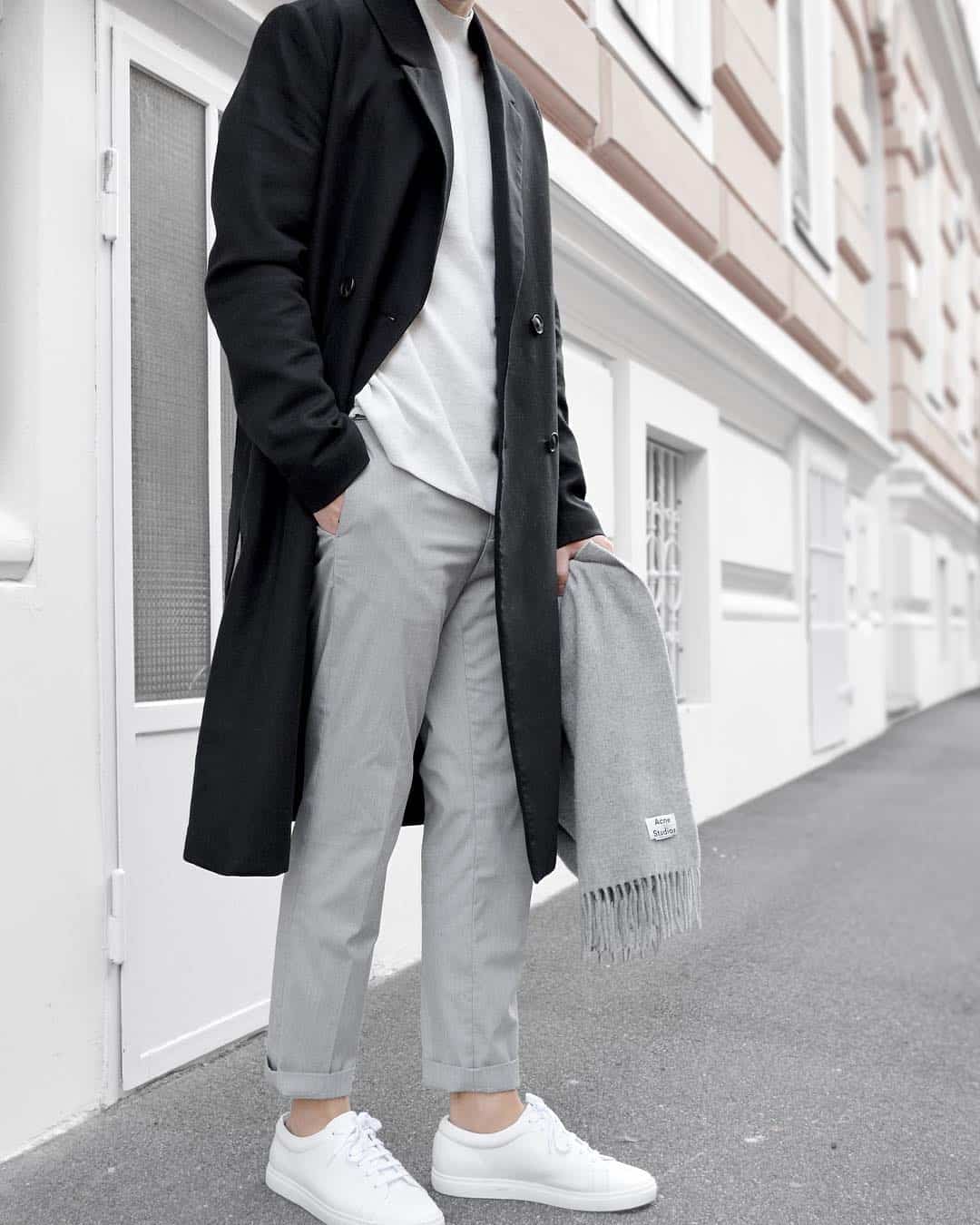 a man wearing grey chinos with a white top, a black trench coat, and white sneakers