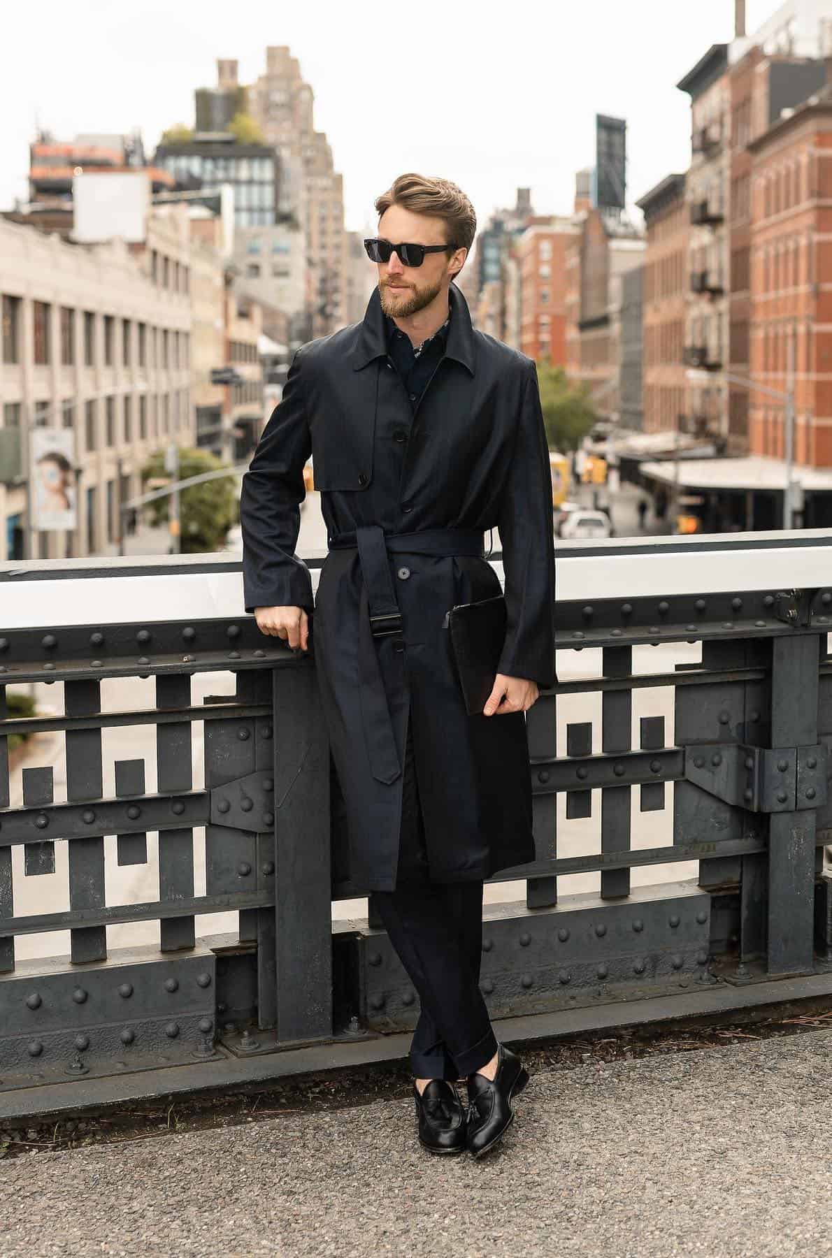 a man wearing black dress pants, black dress shoes, and a black trench coat