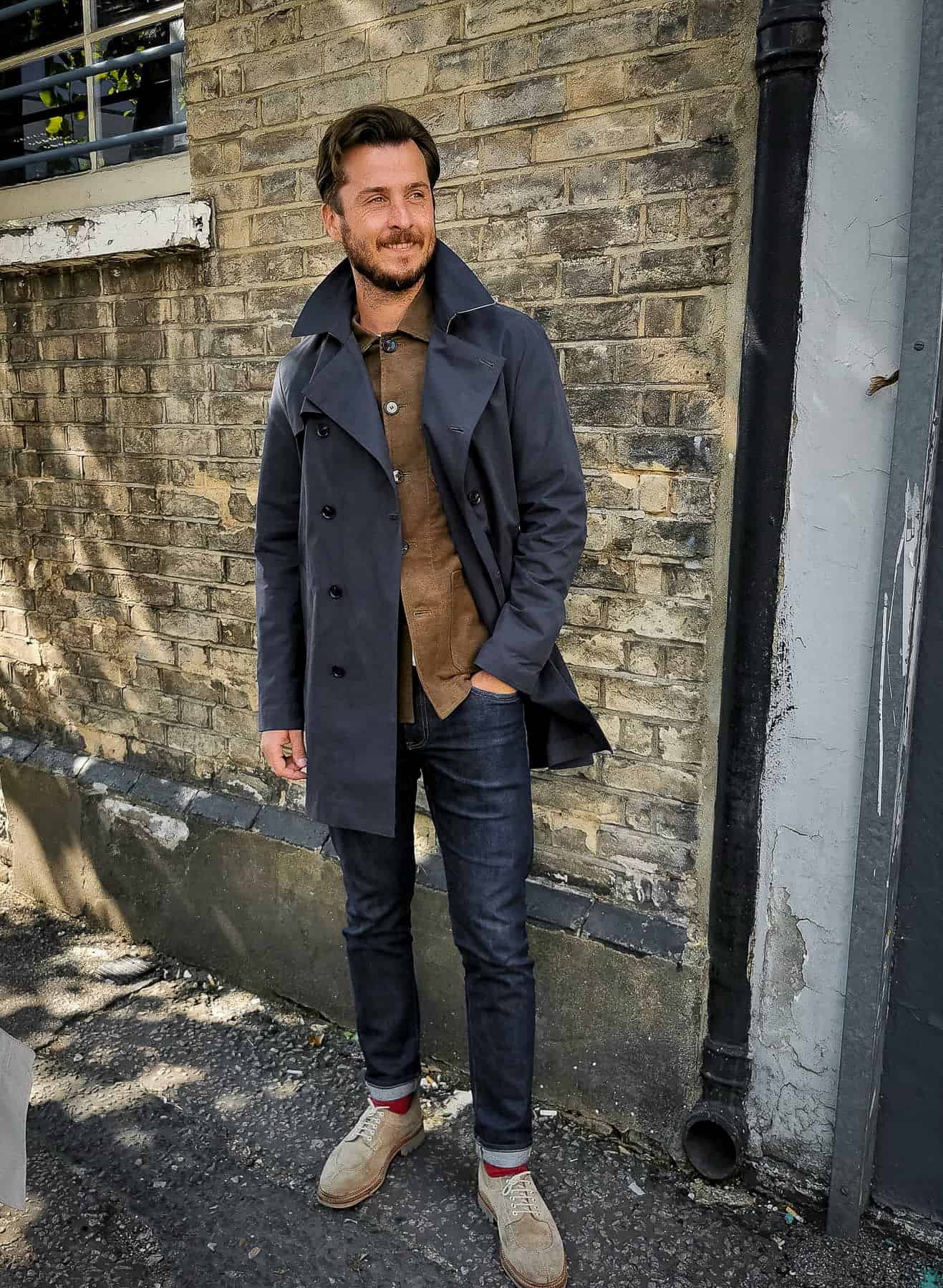 a man wearing dark blue jeans with a brown button-up, a black trench coat, and beige oxford shoes