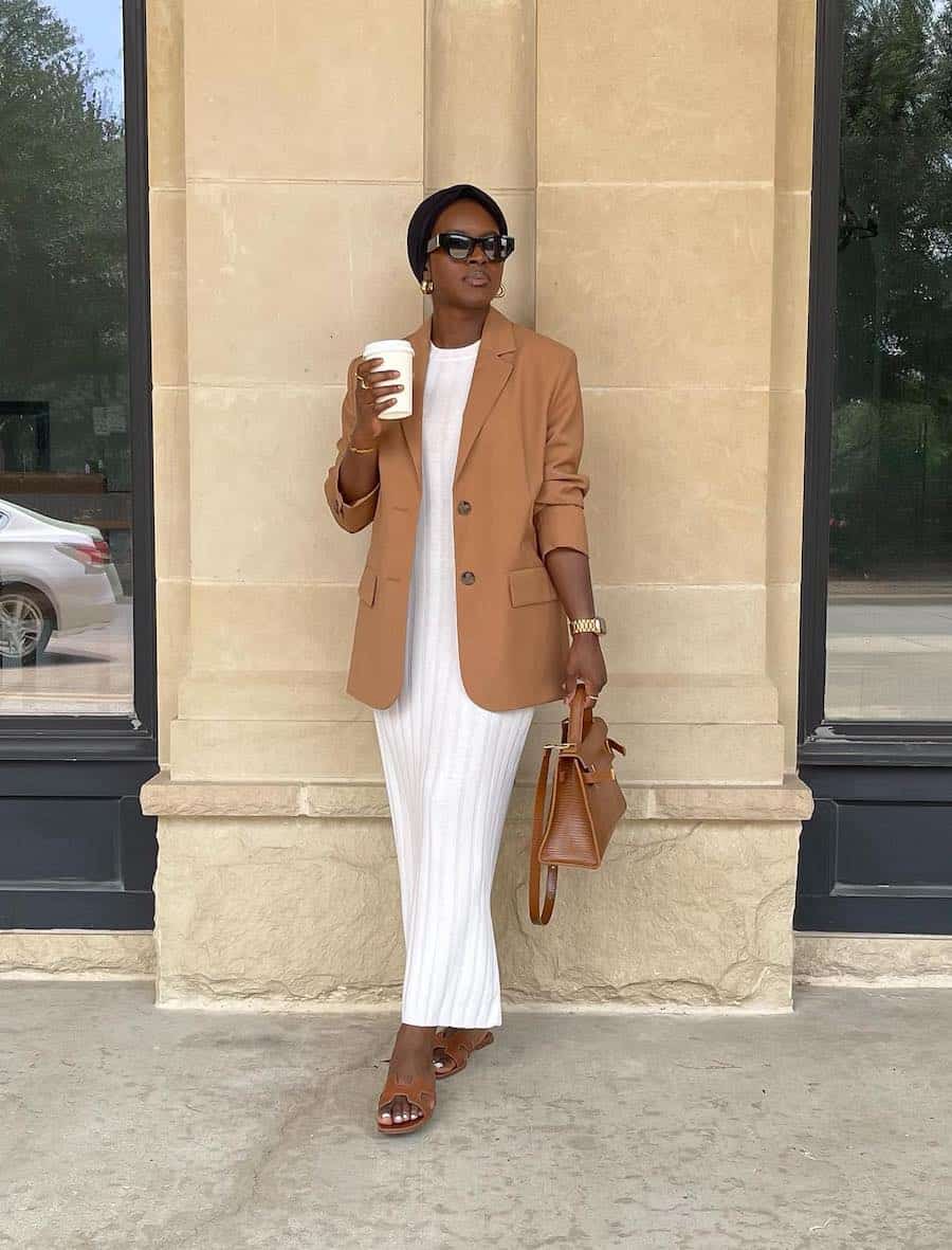 Black woman wearing a camel blazer over an ivory ribbed knit maxi dress with sandals