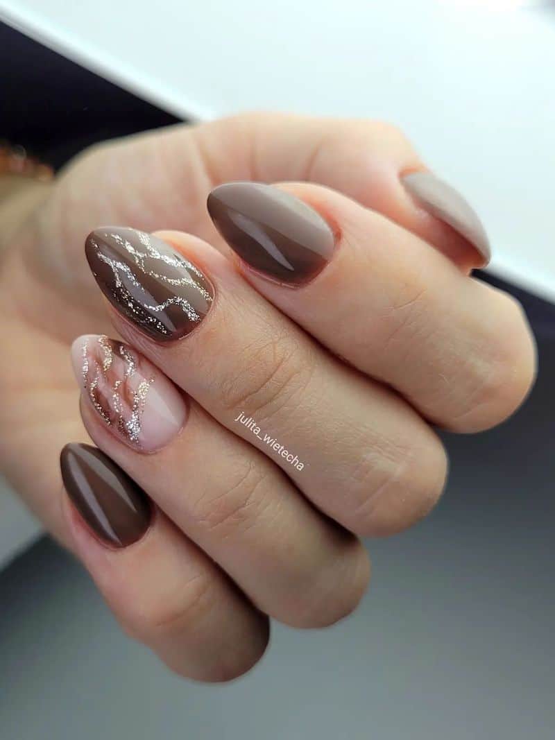 A hand with short brown almond nails with a marbled brown polish accent nails and silver glitter waves