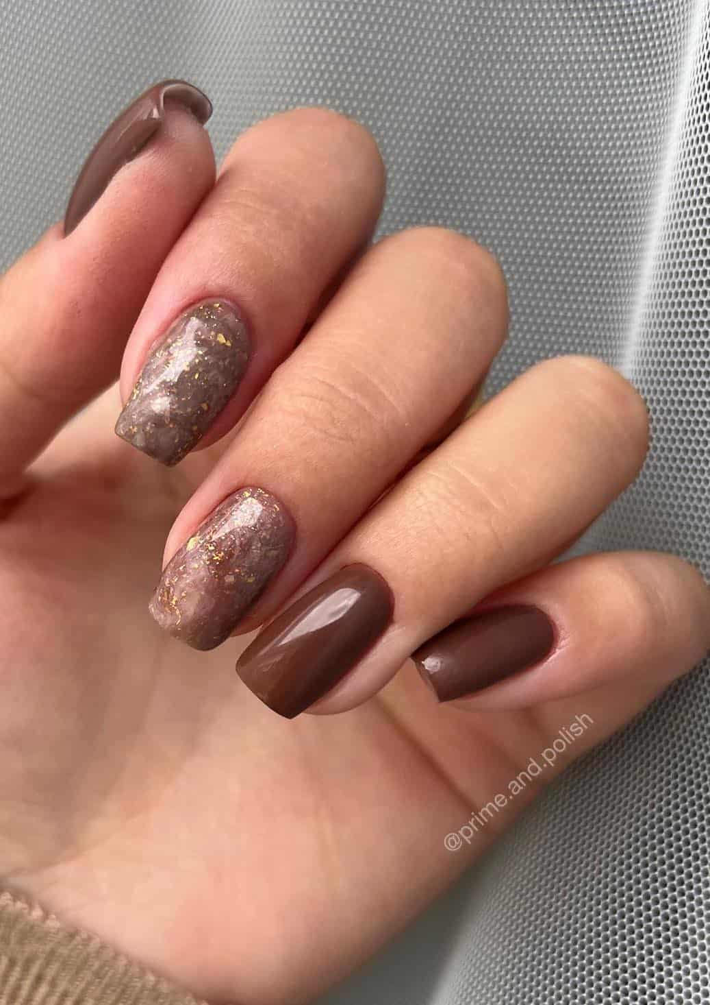 A hand with short square nails painted a glossy brown color with two marbled brown and white accent nails