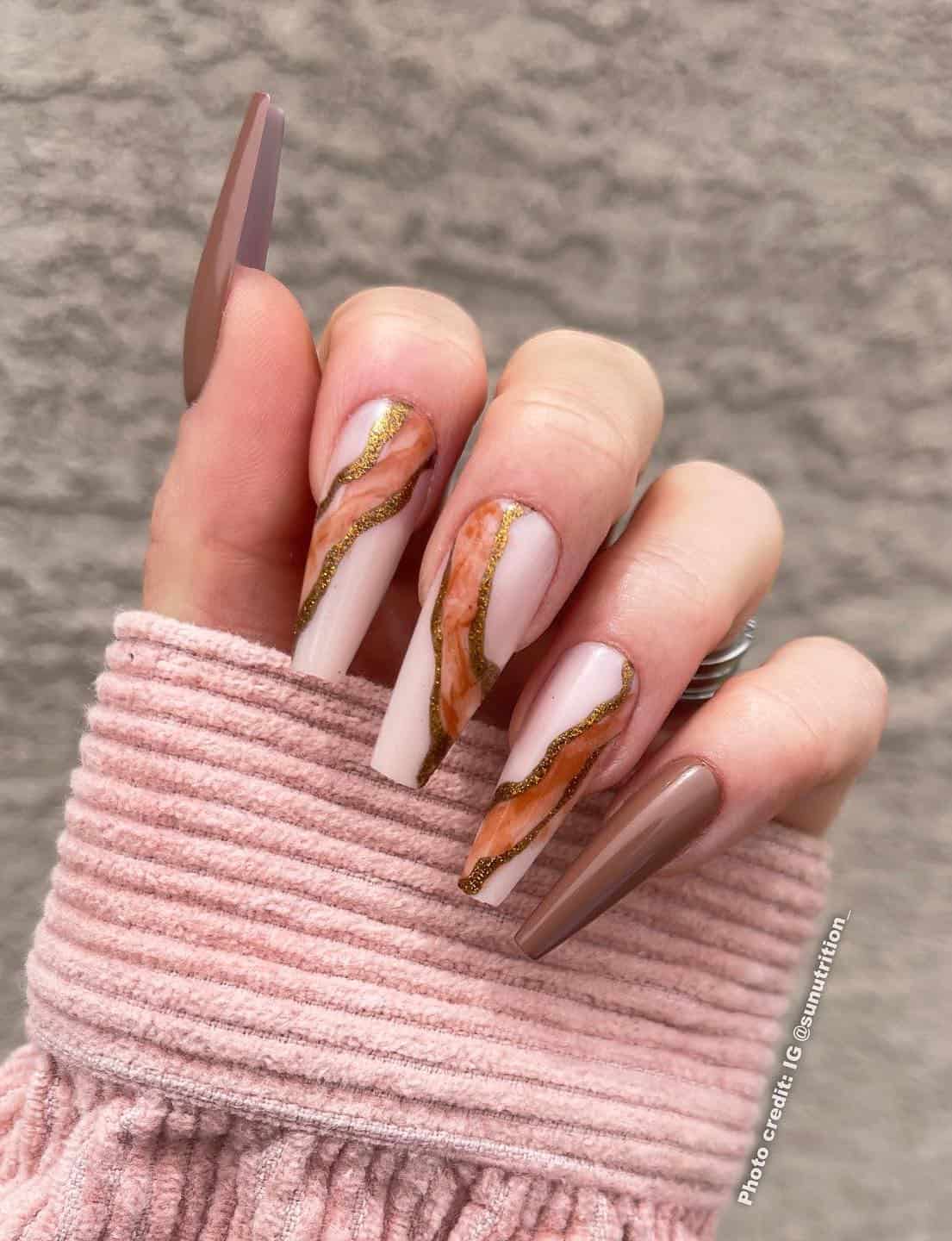 A hand with long coffin nails with two brown nails and three nude pink nails with marbled brown art and gold borders