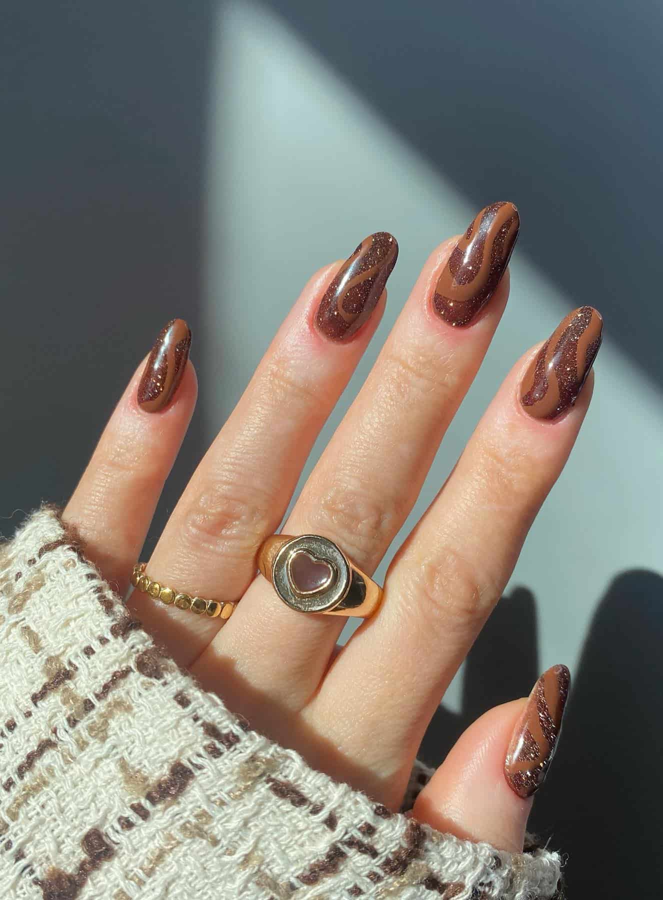 A hand with medium almond nails painted in two shades of brown polish in a swirl design with glitter details