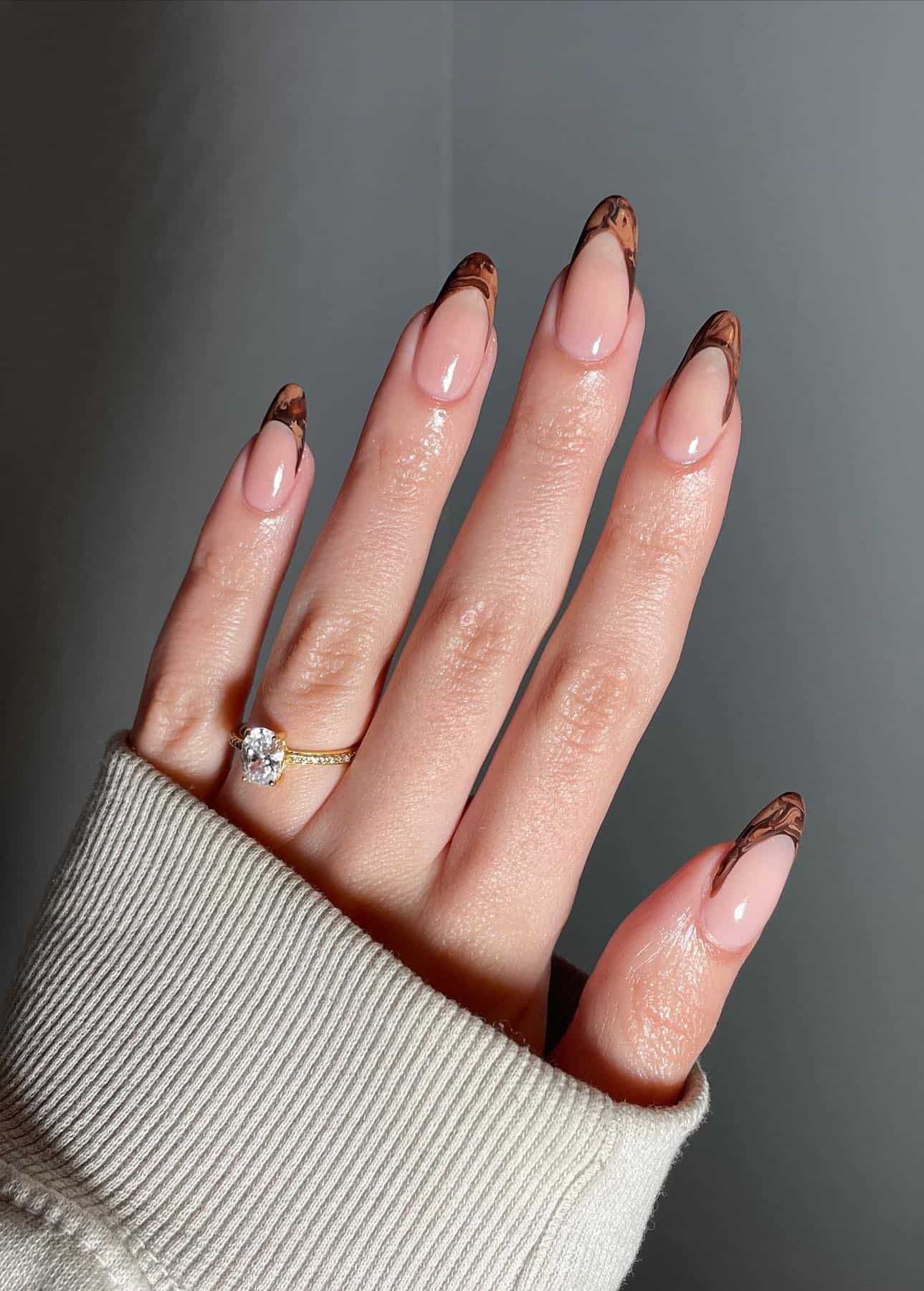 A hand with medium almond nails painted with brown marbled French tips