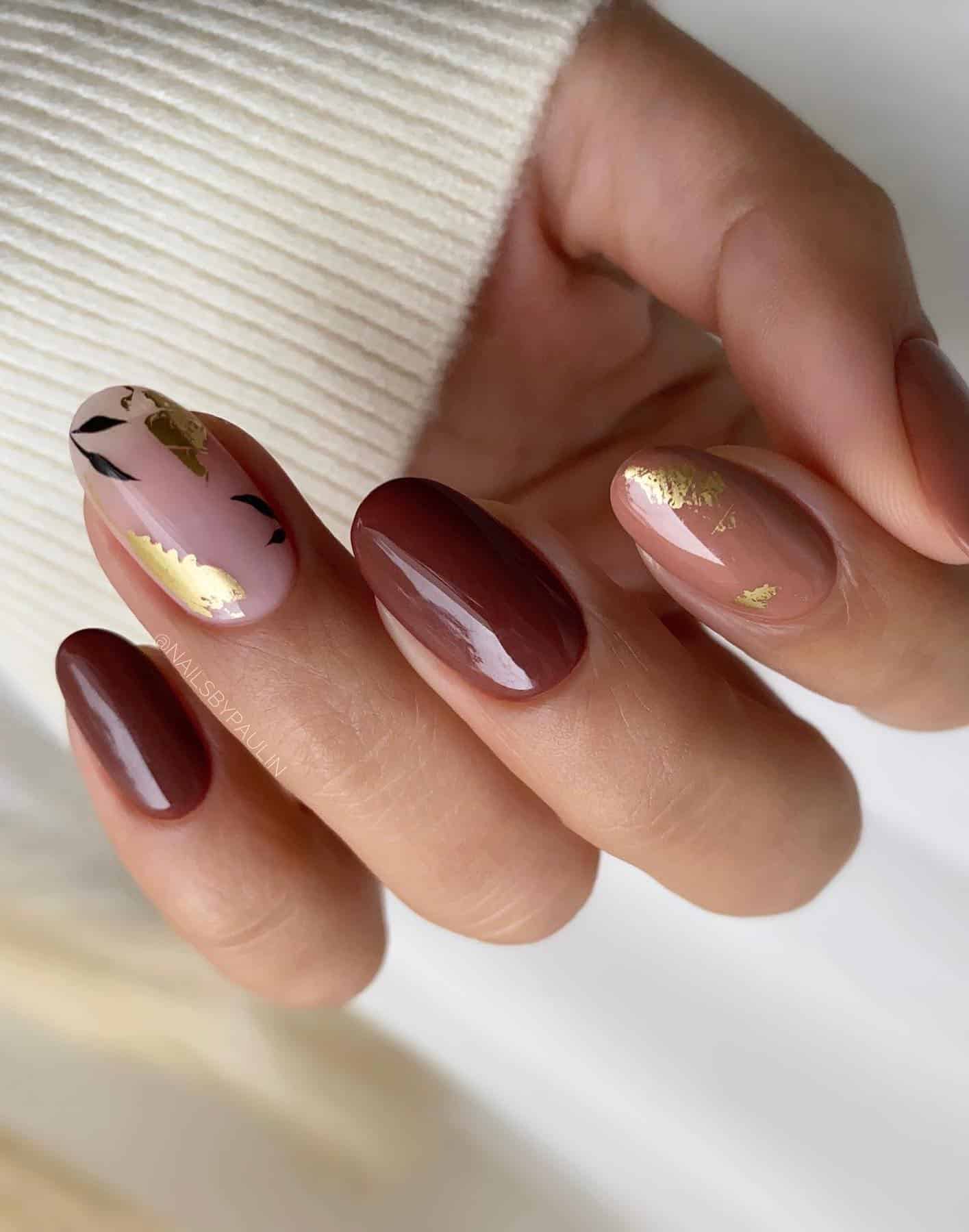 A hand with short round nails painted beige and brown with a nude accent nail with black leaf nail art and gold flake details