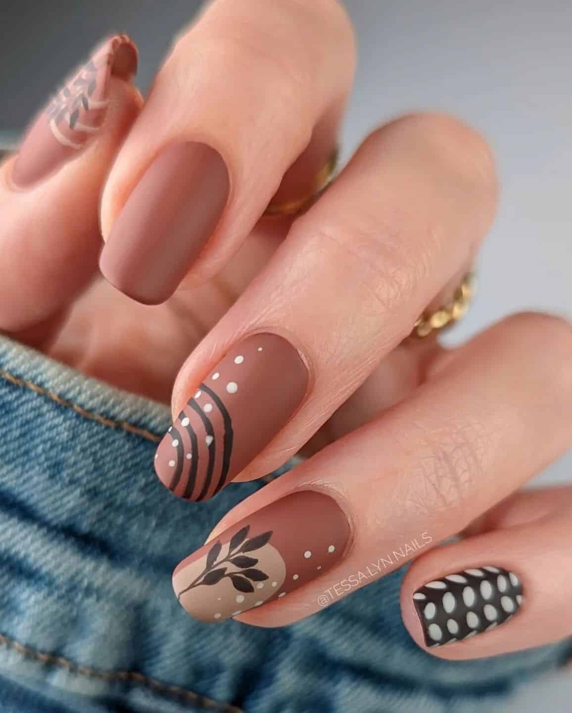 A hand with medium squoval nails painted in a matte brown polish with abstract and leaf nail art