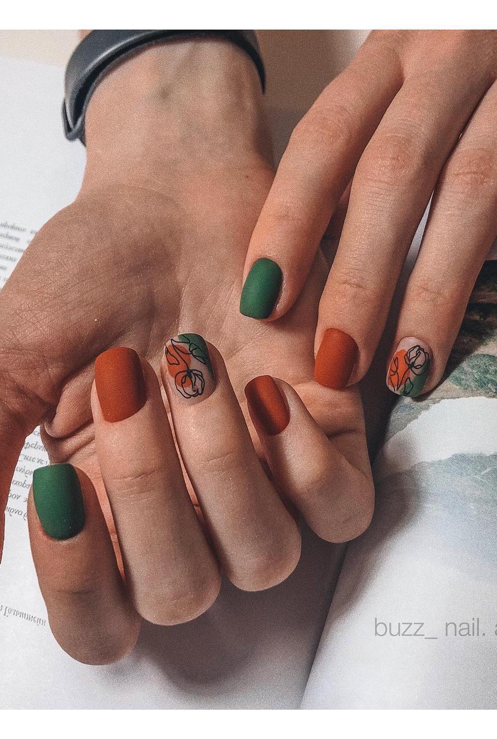 a hand with short square nails painted in matte dark green and burnt orange with a nude accent nail featuring both shades and black floral line art