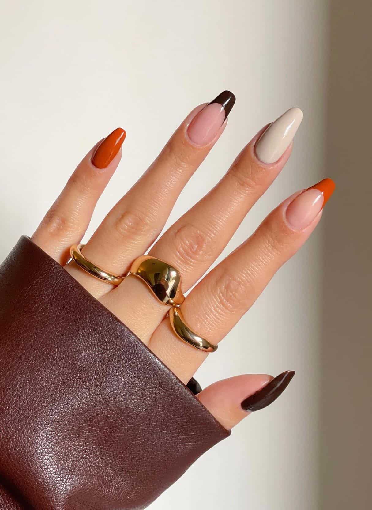 A hand with burnt orange, dark brown, and cream solid-colored nails plus two accent nails with French tips
