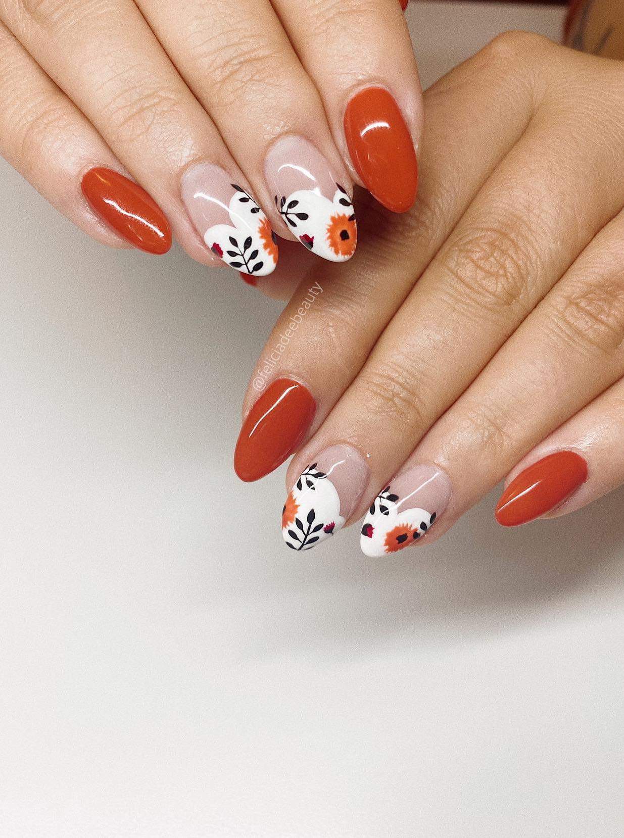 a hand with medium almond nails painted in solid-colored burnt orange and white French tipped accent nails with floral nail art