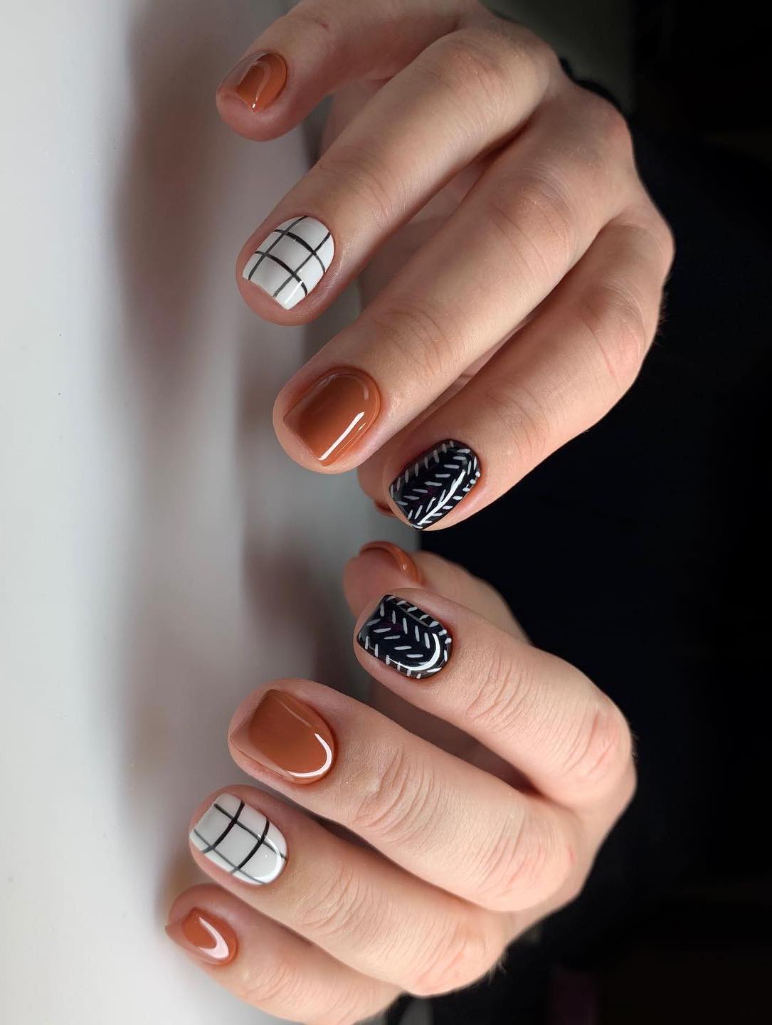 a hand with short square nails painted with solid colored burnt orange, white nails with black grids, and black nails with a white line pattern
