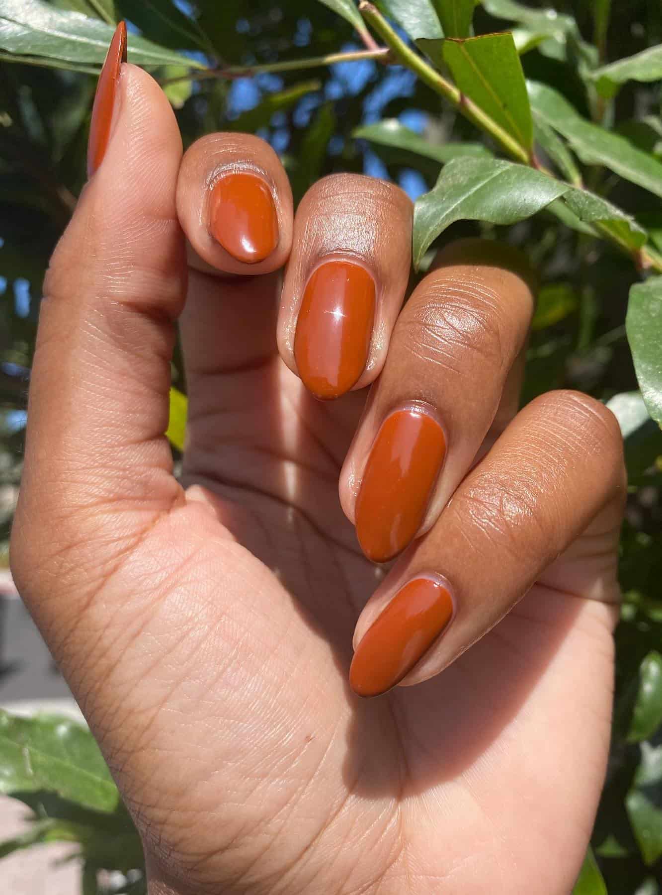 a hand with medium round nails painted with a burnt orange polish and gloss finish