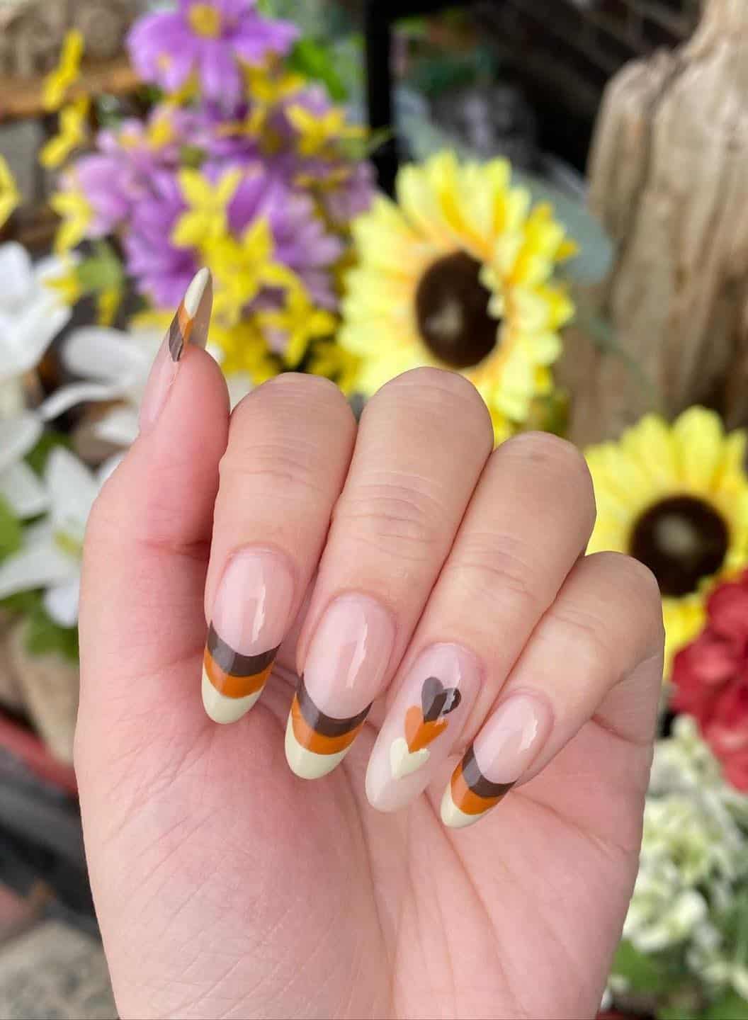 a hand with long round nails painted in cream, burnt orange, and brown French tips with a heart accent nail