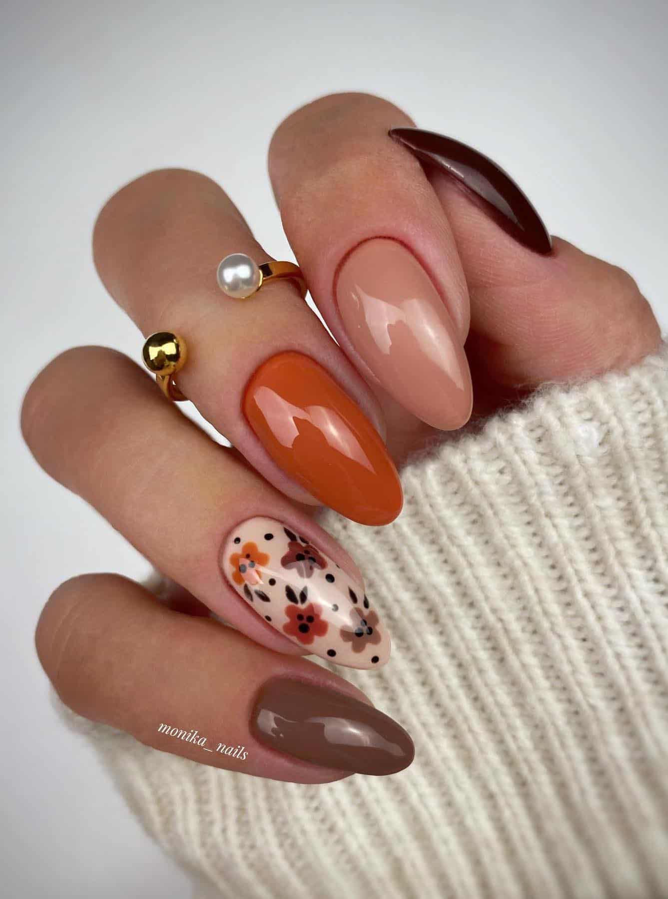 a hand with burnt orange, beige, and brown solid-colored nails plus one cream accent nail with floral art