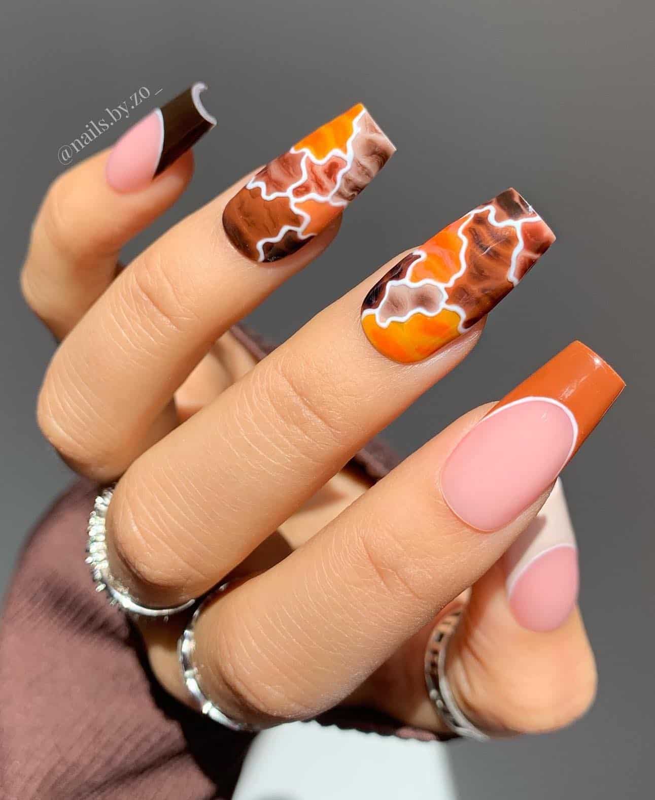 a hand with long coffin nails featuring burnt orange and brown marbled designs with white borders and French tip accent nails