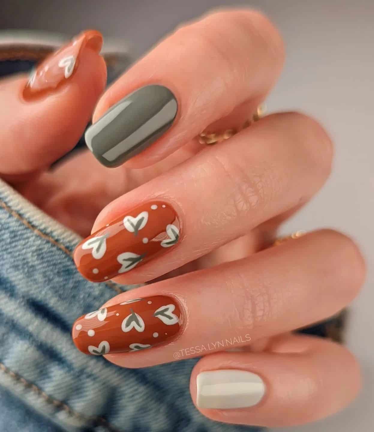 a hand with medium squoval nails painted a burnt orange with leaf nail art and two solid-colored accent nails in white and dark green