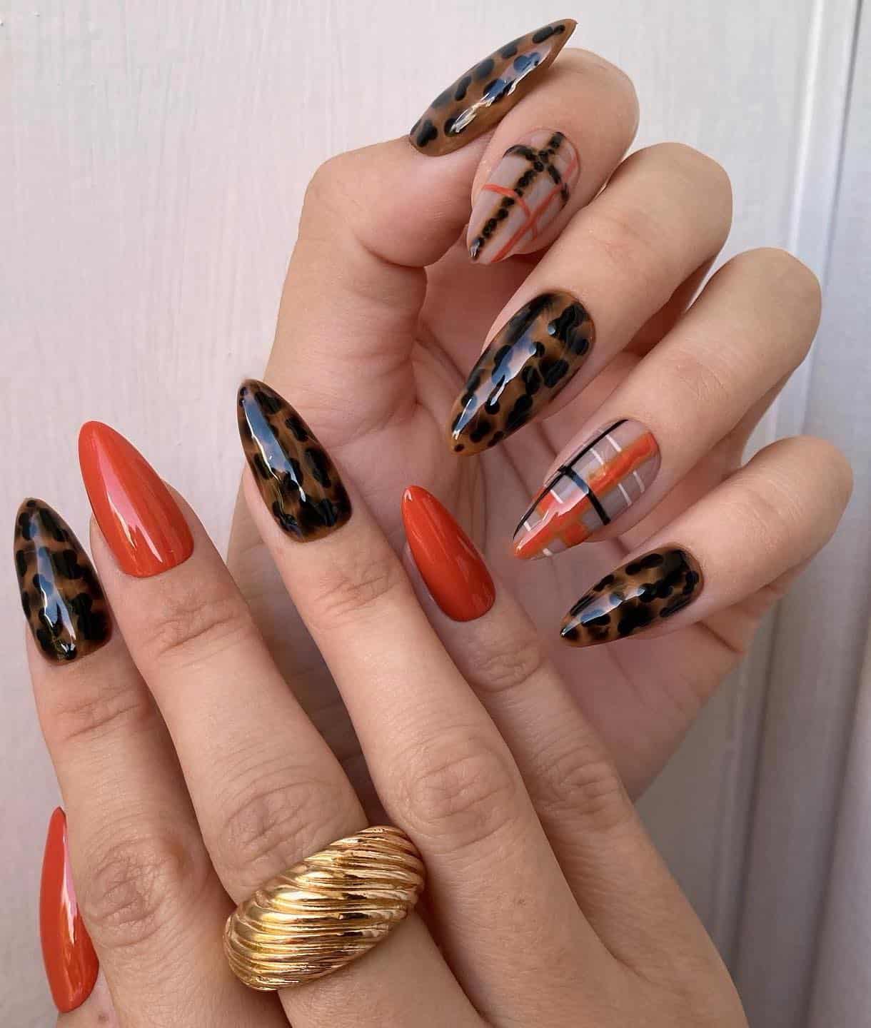 a hand with medium stiletto nails featuring multiple designs including solid colored burnt orange nails, black and brown tortoise shell nails, and black, white, and orange plaid nails