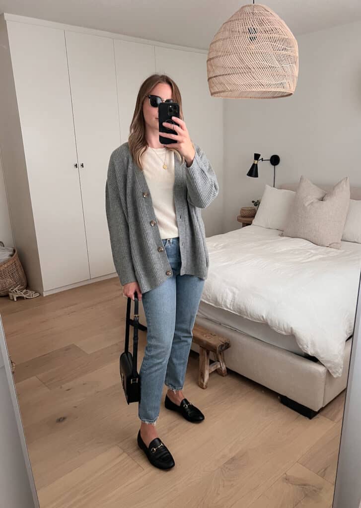 woman wearing a fall capsule wardrobe outfit with a grey knit cardigan over a knit ivory tee, jeans, and black loafers