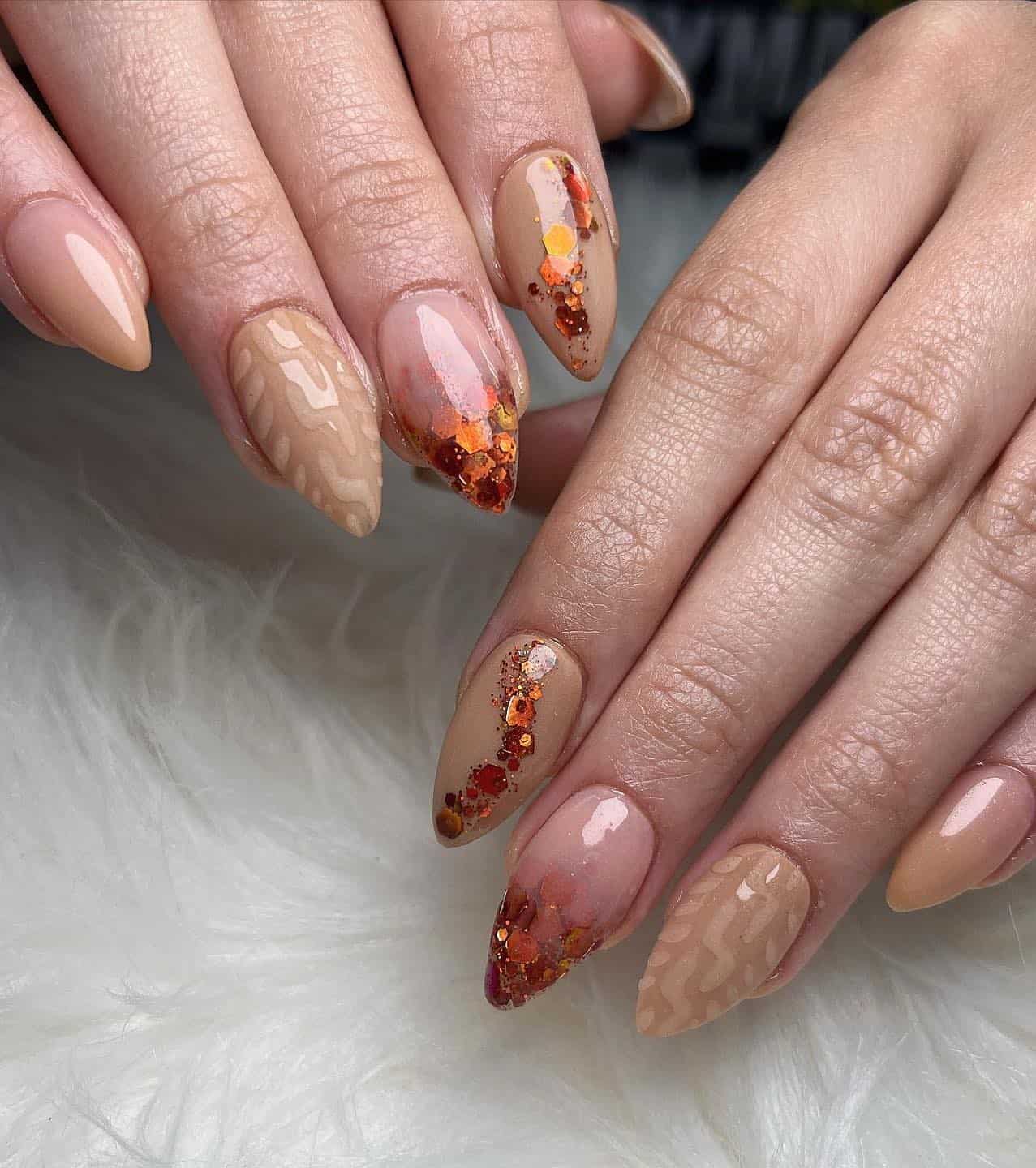 a hand with short stiletto nails painted in multiple designs, including beige sweater nails, nude ad beige ombre, and nude and orange glitter ombre