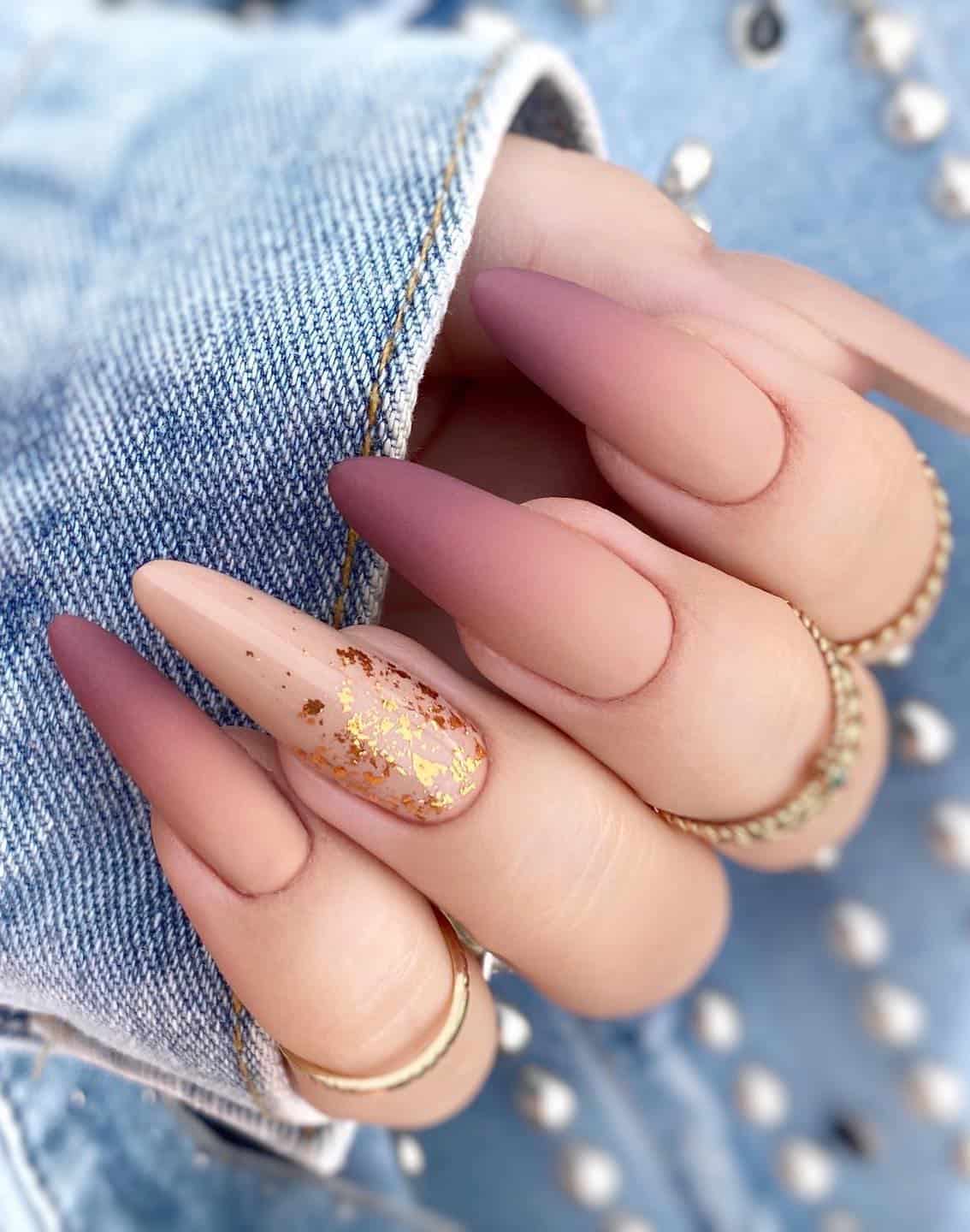 a hand with long almond nails painted a matte beige and purple ombre with a glossy beige accent nail featuring gold flake details