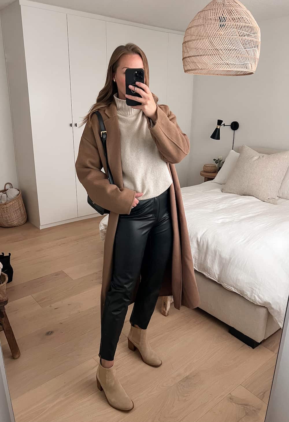 woman wearing a long camel coat over a beige turtleneck sweater with leather pants and tan suede ankle boots