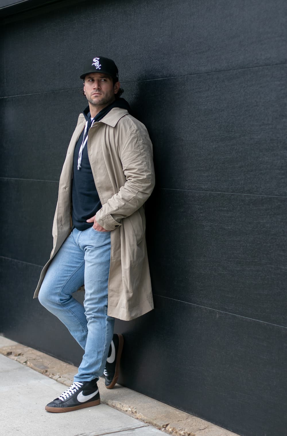 a man wearing light blue jeans, a hoodie, a beige trench coat, and sneakers
