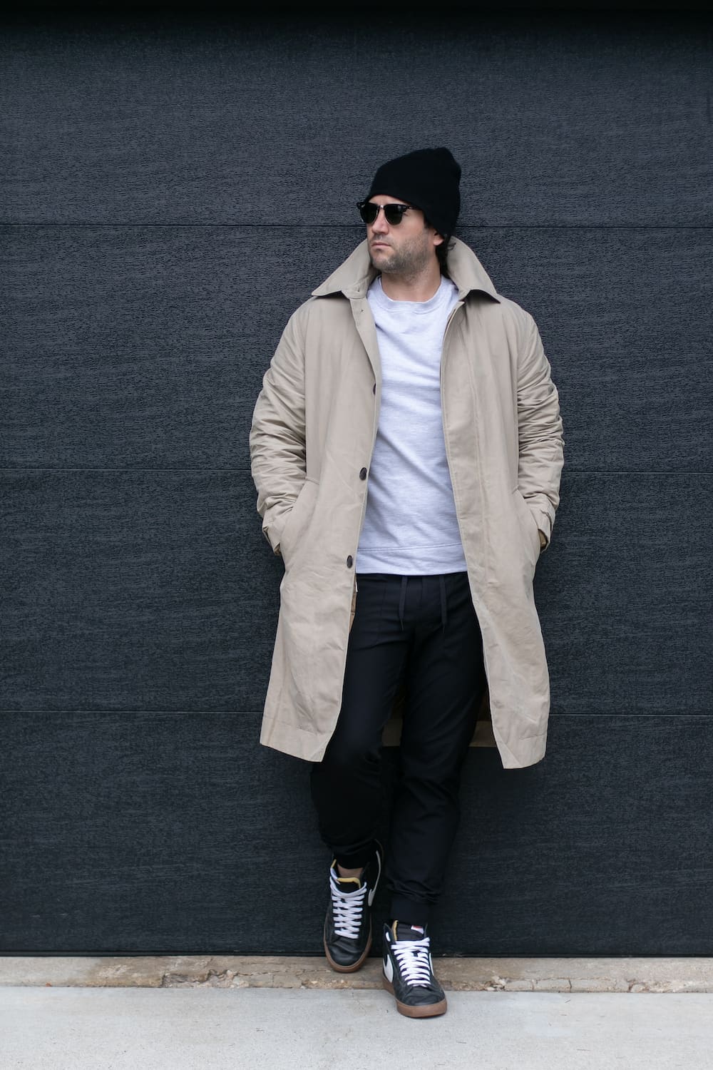 a man wearing black joggers with a white tee, a beige trench coat, black sneakers, and a beanie
