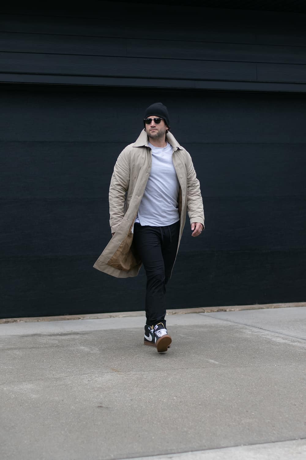 a man wearing black joggers with a white tee, a beige trench coat, black sneakers, and a beanie